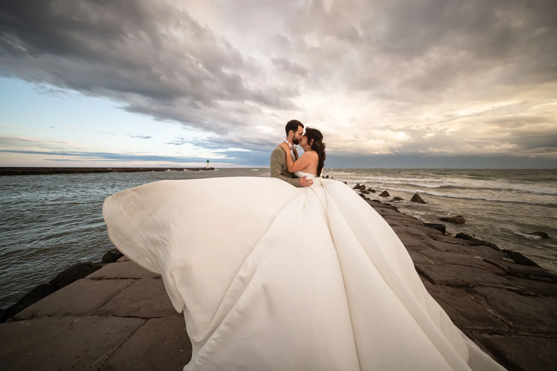 Séance photo couple après mariage à Agde | Studio Graou Mariés s’embrassant au bord de la mer à Agde, avec la robe de mariée déployée sous un ciel nuageux.